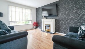 A modern living room with a fireplace, a flat-screen TV mounted above it, and a patterned wallpaper. - Livingston 4 Bed + Car Space (West Lothian)
