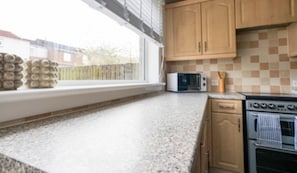 A kitchen with wooden cabinets, a microwave, an oven, and a window with blinds. - Livingston 4 Bed + Car Space (West Lothian)