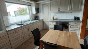 Uma cozinha compacta com uma mesa de madeira, cadeiras pretas, armários brancos, micro-ondas e uma janela com flores cor-de-rosa do lado de fora.
