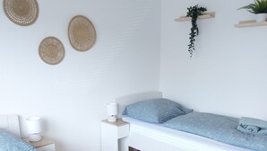 Quarto moderno com duas camas, uma mesinha de cabeceira com abajur, um tapete pequeno e decorações nas paredes, incluindo cestos de palha e uma planta pendente.
