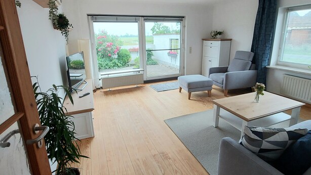 Uma sala de estar moderna com piso de madeira, sofá cinza e uma mesa de jantar. Há uma porta de vidro deslizante que dá para uma varanda com plantas e flores.