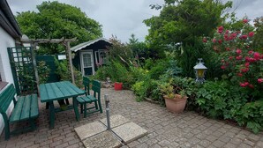 Um pátio com jardim, mesa, cadeiras, um pequeno depósito e diversas plantas.