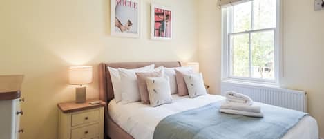 Un dormitorio con cama, mesitas de noche, lámparas, un ventanal con vistas y dos cuadros en la pared.