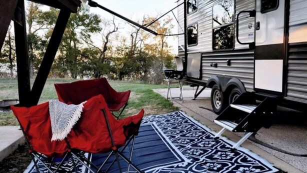 Een camper met een overdekte veranda, een barbecue en een rode opvouwbare stoel.
