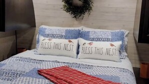 Een bed met een deken met een blauw-wit patroon, twee kussens met de tekst 'BLESSS THIS NEST.', een rode plaid en een aan de muur bevestigd krans.