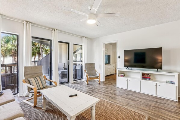A spacious living area with a white ceiling fan, a flat-screen TV on a white cabinet, and a sliding glass door leading to an outdoor patio.