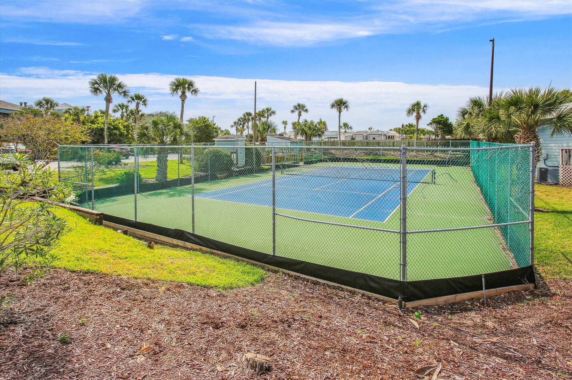 Un court de tennis entouré d’une clôture en treillis métallique, avec une pelouse et des arbres en arrière-plan.