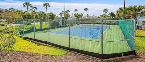 Un court de tennis entouré d’une clôture en treillis métallique, avec une pelouse et des arbres en arrière-plan.
