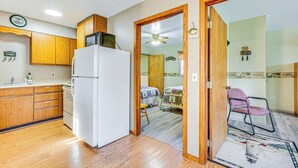 A compact kitchen with wooden cabinets, a white refrigerator, and a microwave on top. A wooden door leads to a bedroom with a bed and a chair. - Michigan Manistee River Cabin ~ 1 Mi to Downtown (Manistee)