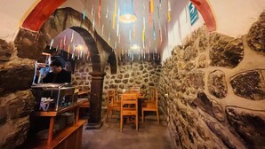 Une pièce aux murs de pierre, avec une table et des chaises en bois, un petit coin bar et des décorations colorées suspendues.