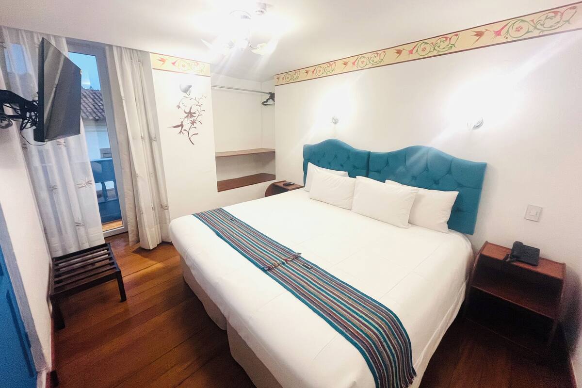 Une chambre d’hôtel avec un grand lit, un plancher en bois, un téléviseur fixé au mur et une fenêtre avec des rideaux.