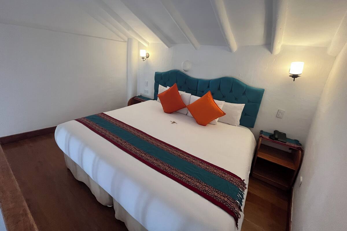 Un lit bien fait, avec une couette blanche, une tête de lit turquoise et des oreillers orange. On y trouve deux tables de chevet avec des lampes, une table de nuit en bois et un luminaire fixé au mur.