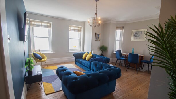 Un salon avec un canapé bleu, un fauteuil jaune et un coin repas avec des chaises bleues.