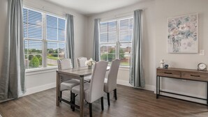 Un comedor con una mesa y sillas de madera, ventanas amplias con persianas, una consola con cajones y un cuadro en la pared.