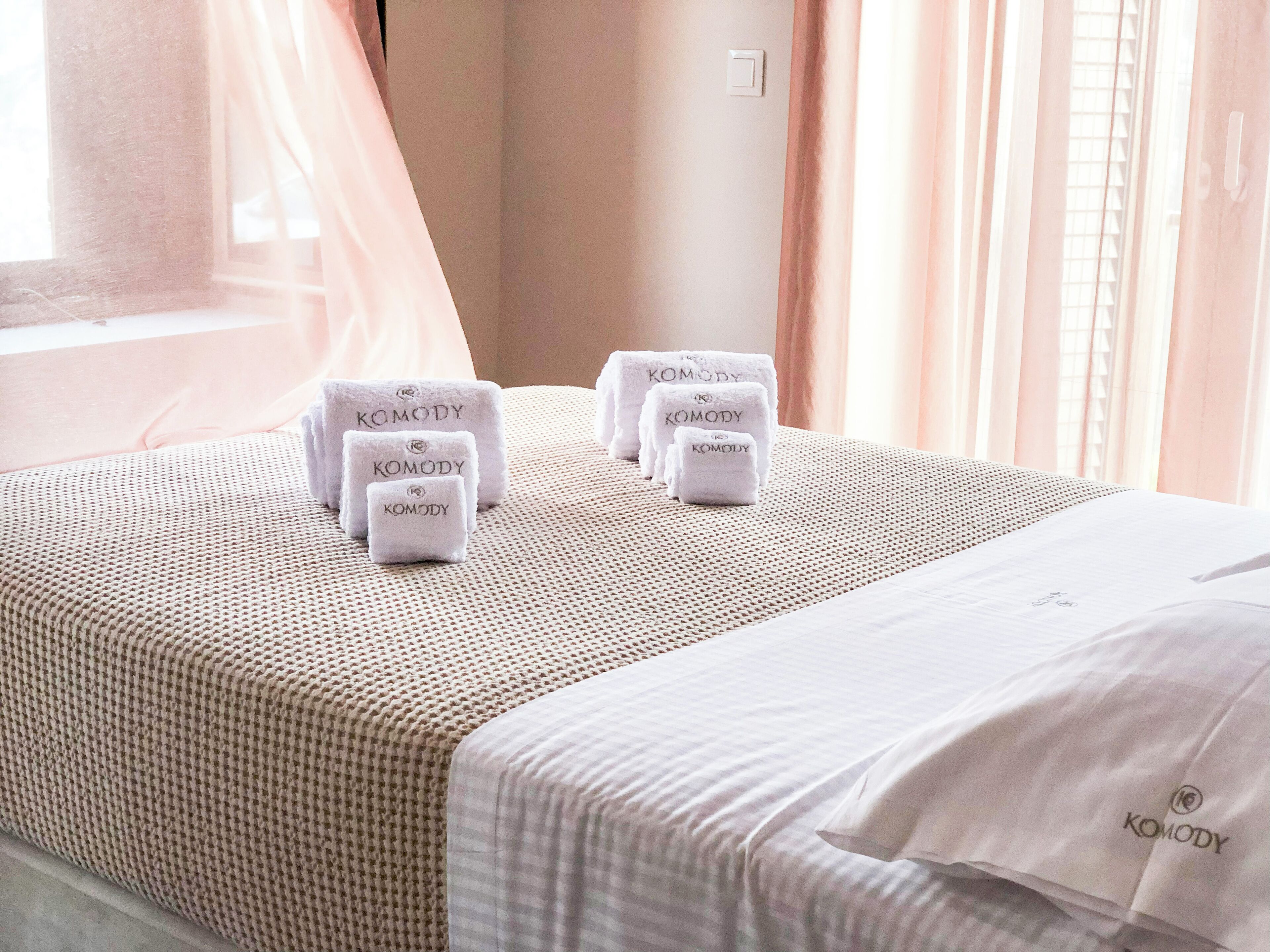 A hotel room with a neatly made bed, a window with sheer curtains, and several towels with the brand name "Komody" on them.