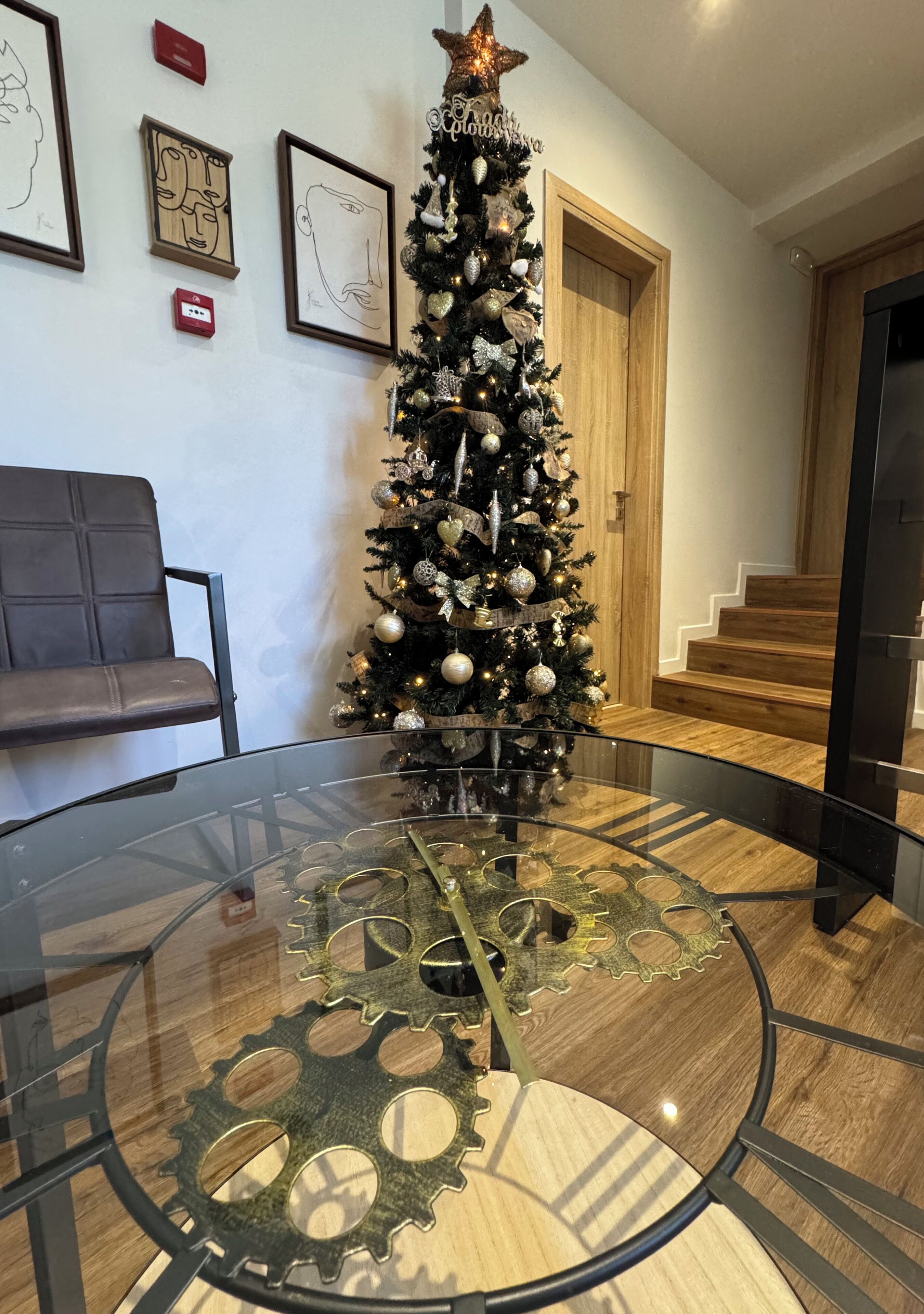 A modern interior with a glass table featuring metal gears, a Christmas tree, framed artwork on the wall, and a wooden bench.