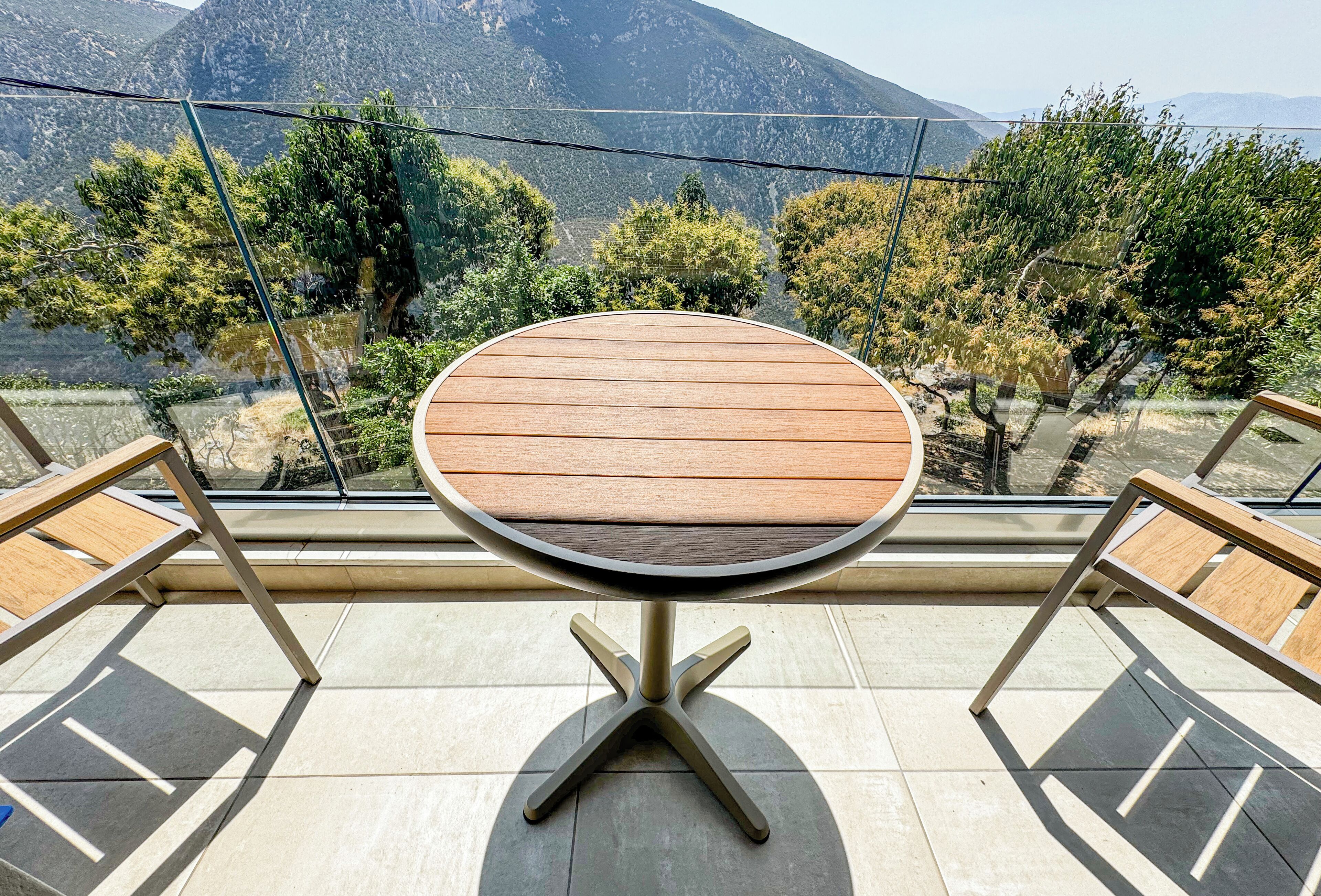 A round table with a wooden top and metal legs, set on a balcony with a view of mountains and trees.