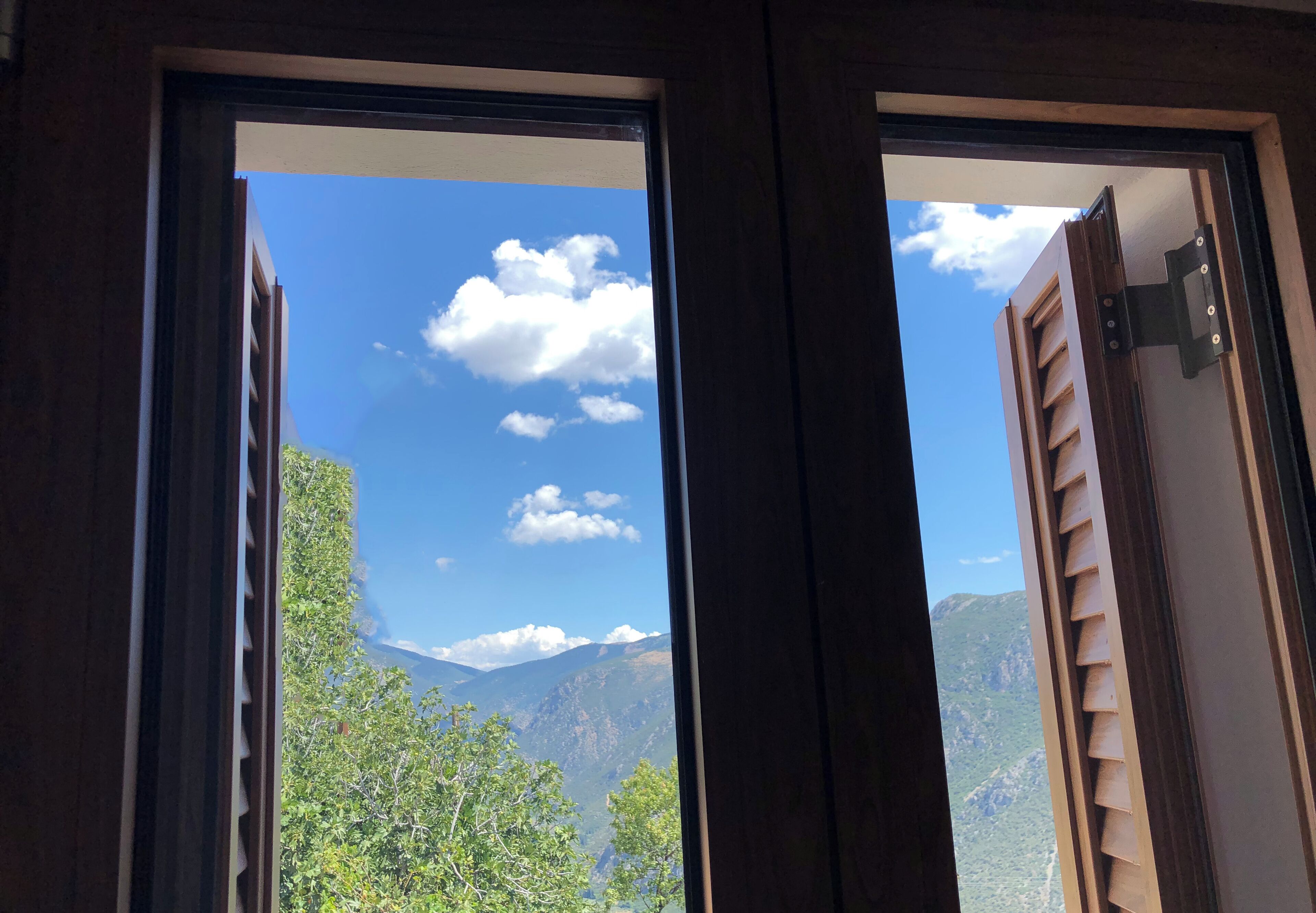 A view through a window with wooden frames, revealing a mountainous landscape with greenery and a clear sky.