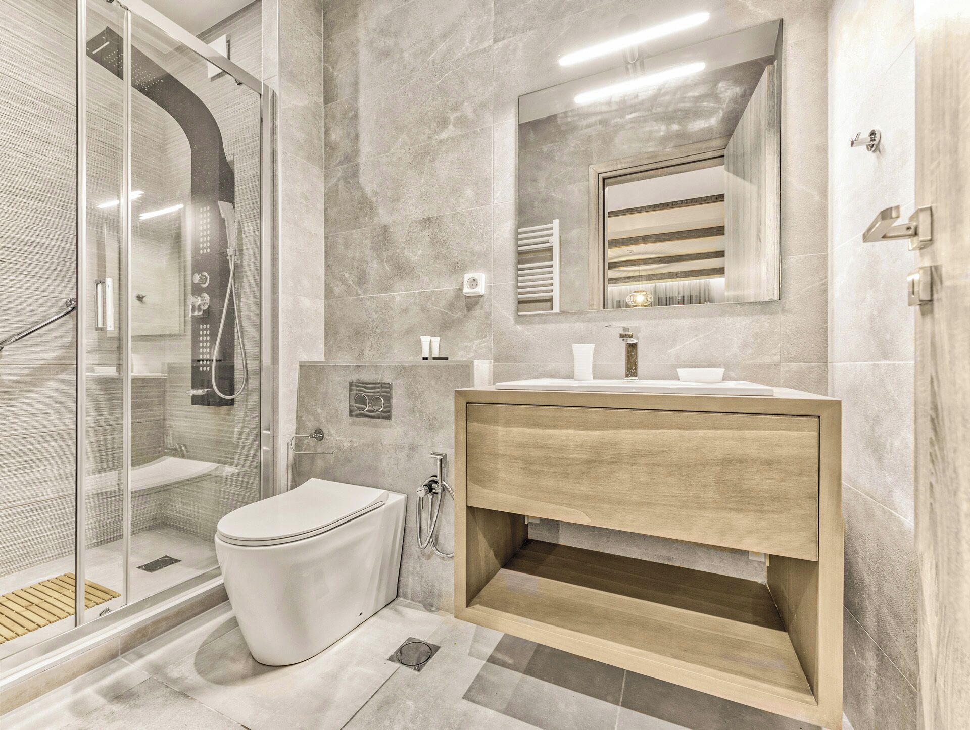 Un baño moderno con ducha envuelta en vidrio, inodoro blanco, tocador de madera y un espejo grande.