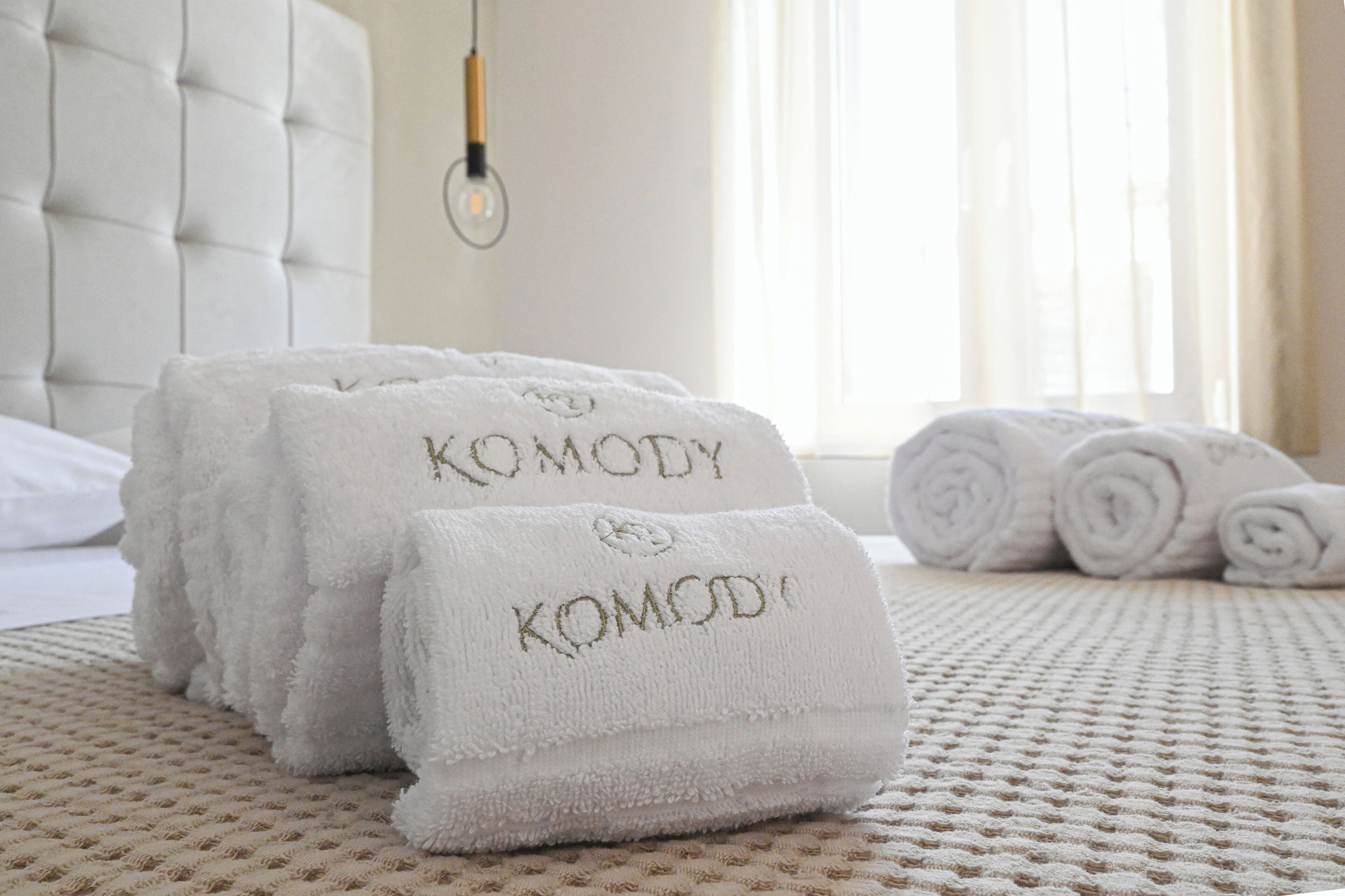 A neatly arranged stack of white towels with embroidered text, a tufted headboard, and a window with curtains in a hotel room.