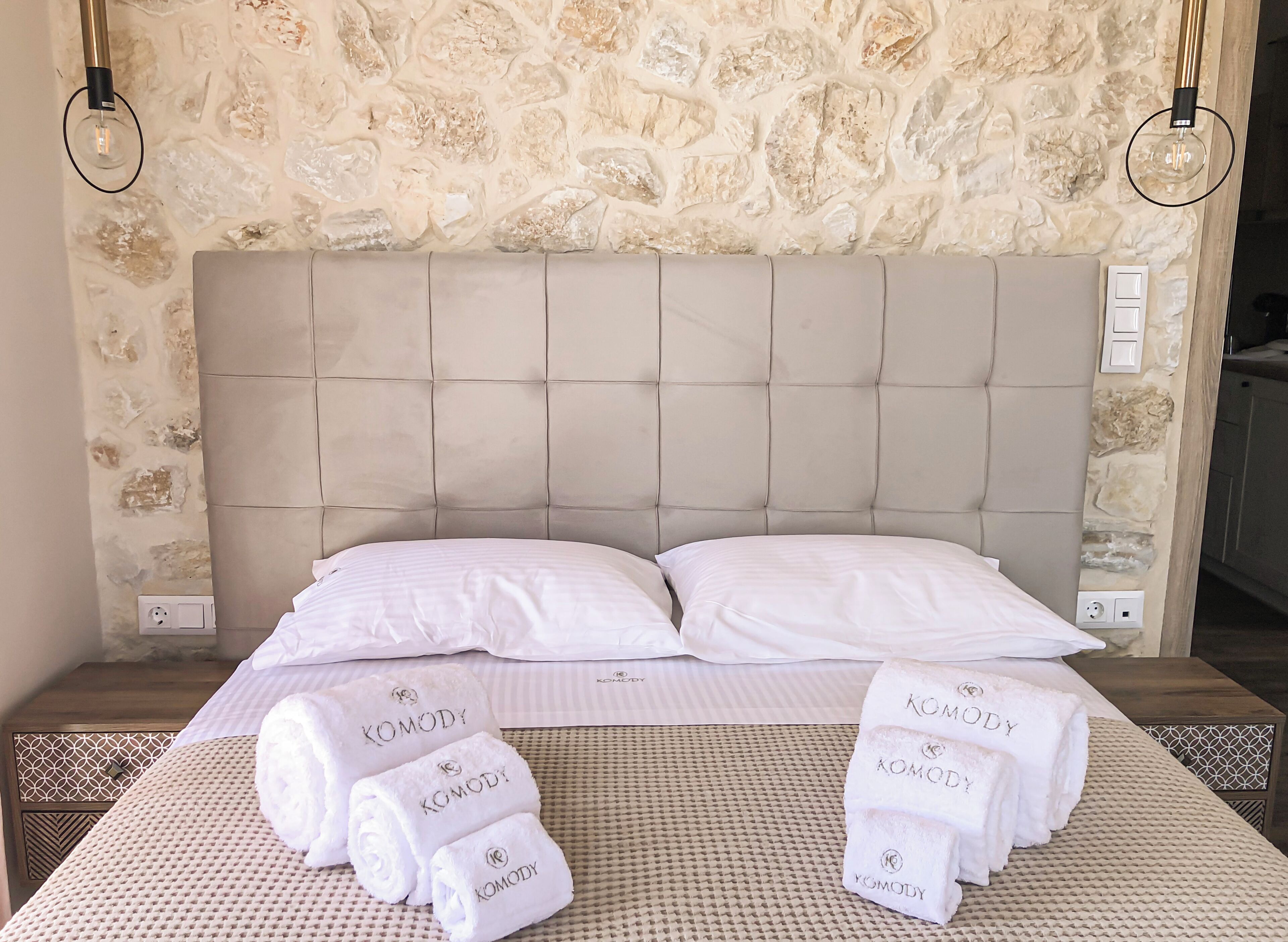 A hotel room with a stone wall, a tufted headboard, two white pillows, and a bed with a woven bedspread.