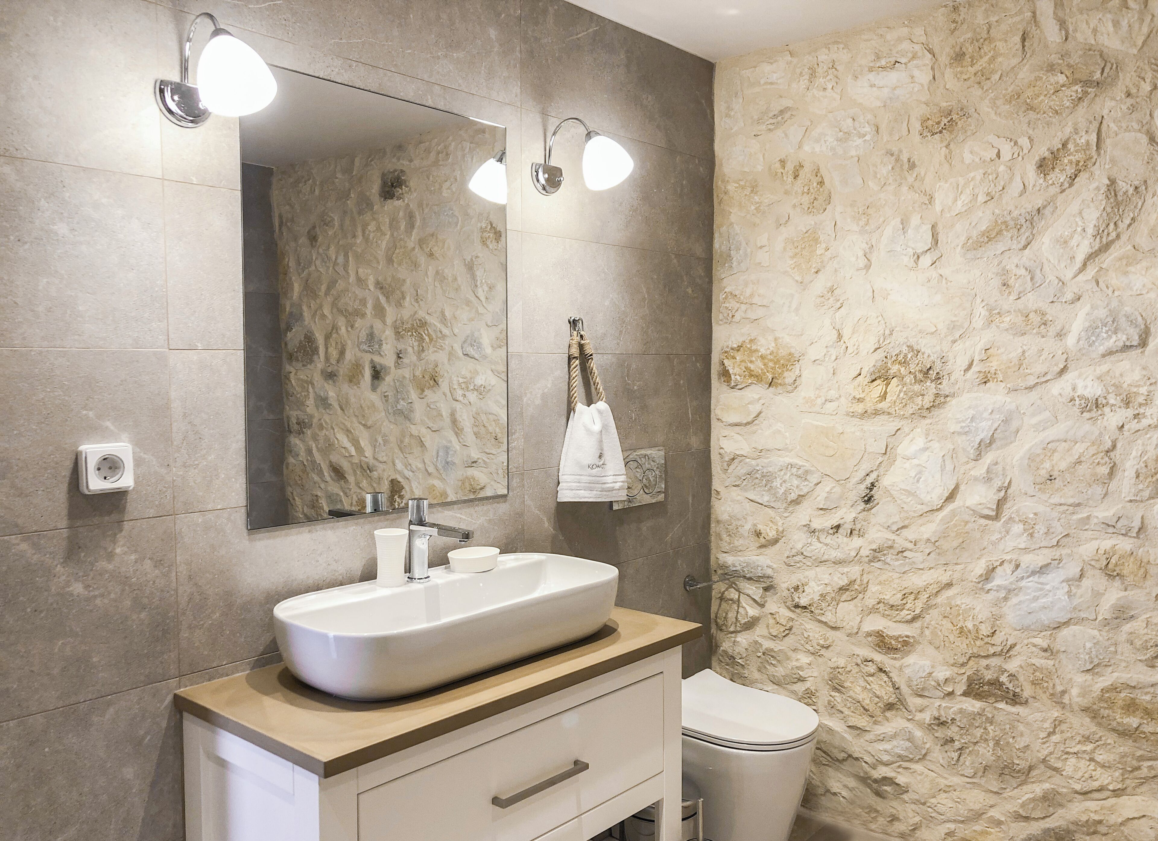 A bathroom with a stone wall, a large mirror, a sink with a towel, and a toilet.