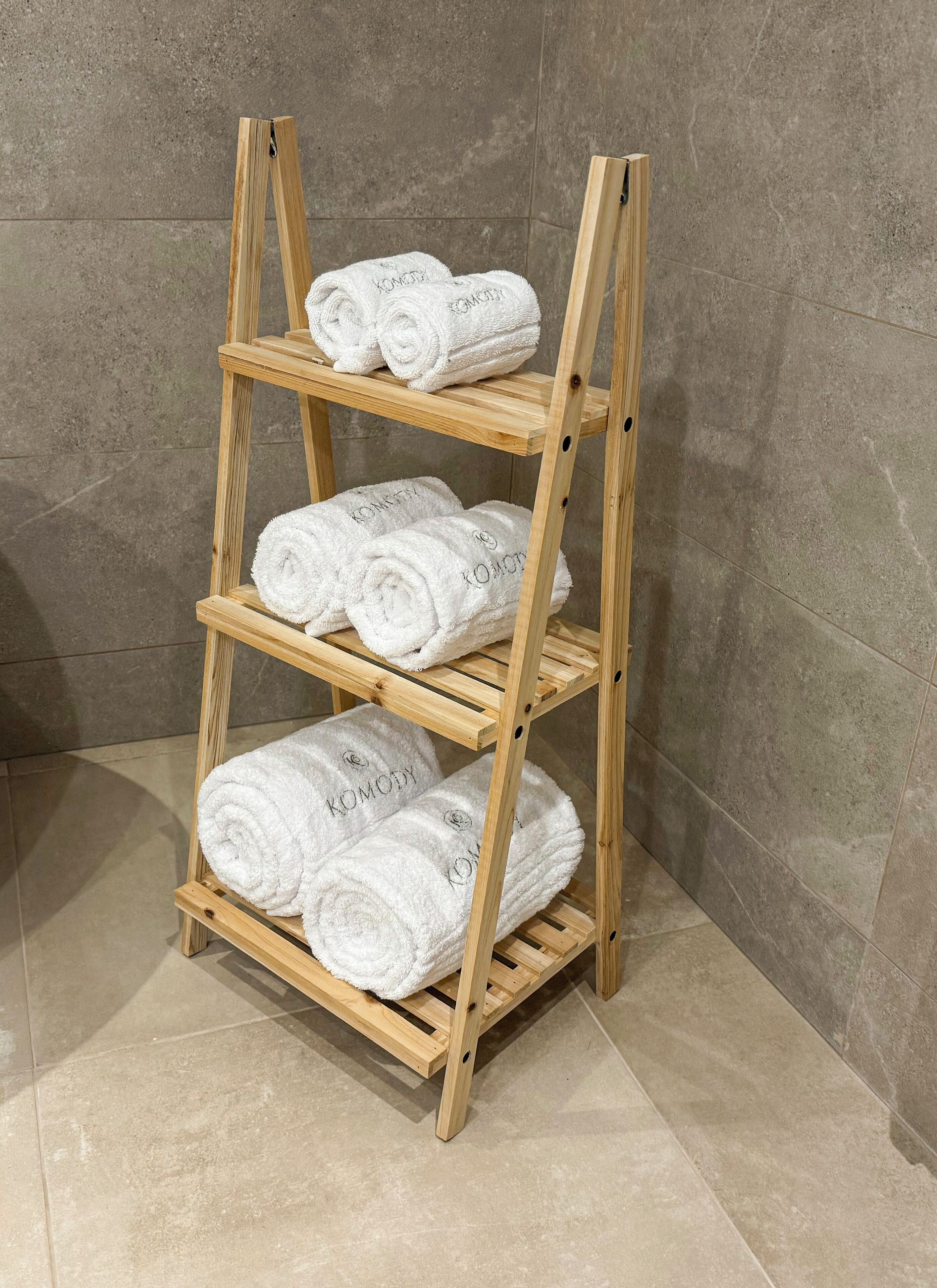 Une étagère en bois à trois niveaux, sur laquelle sont pliées et empilées des serviettes blanches portant l’inscription « KOMODY ».
