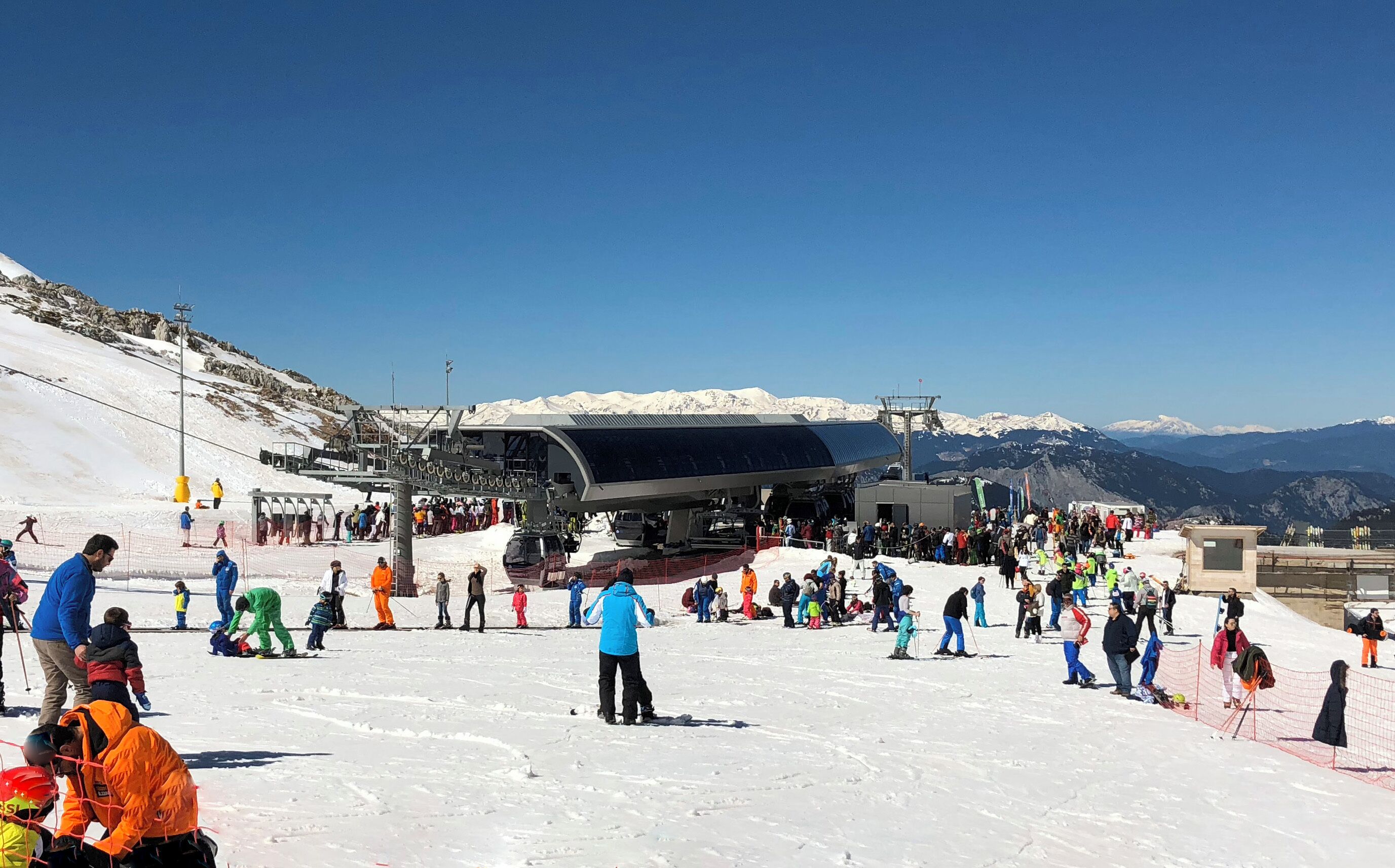 A ski lift with people waiting and skiing on a snowy slope.