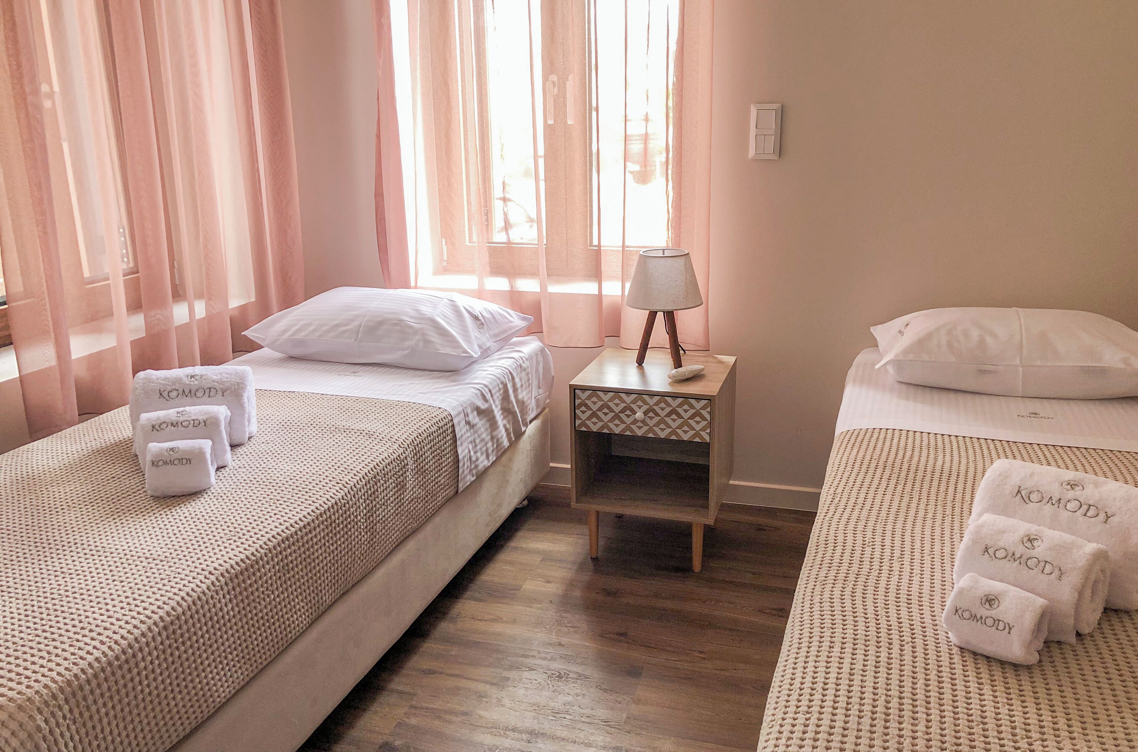 A hotel room with two beds, each with a white bedspread and pillows, and a small bedside table with a lamp.
