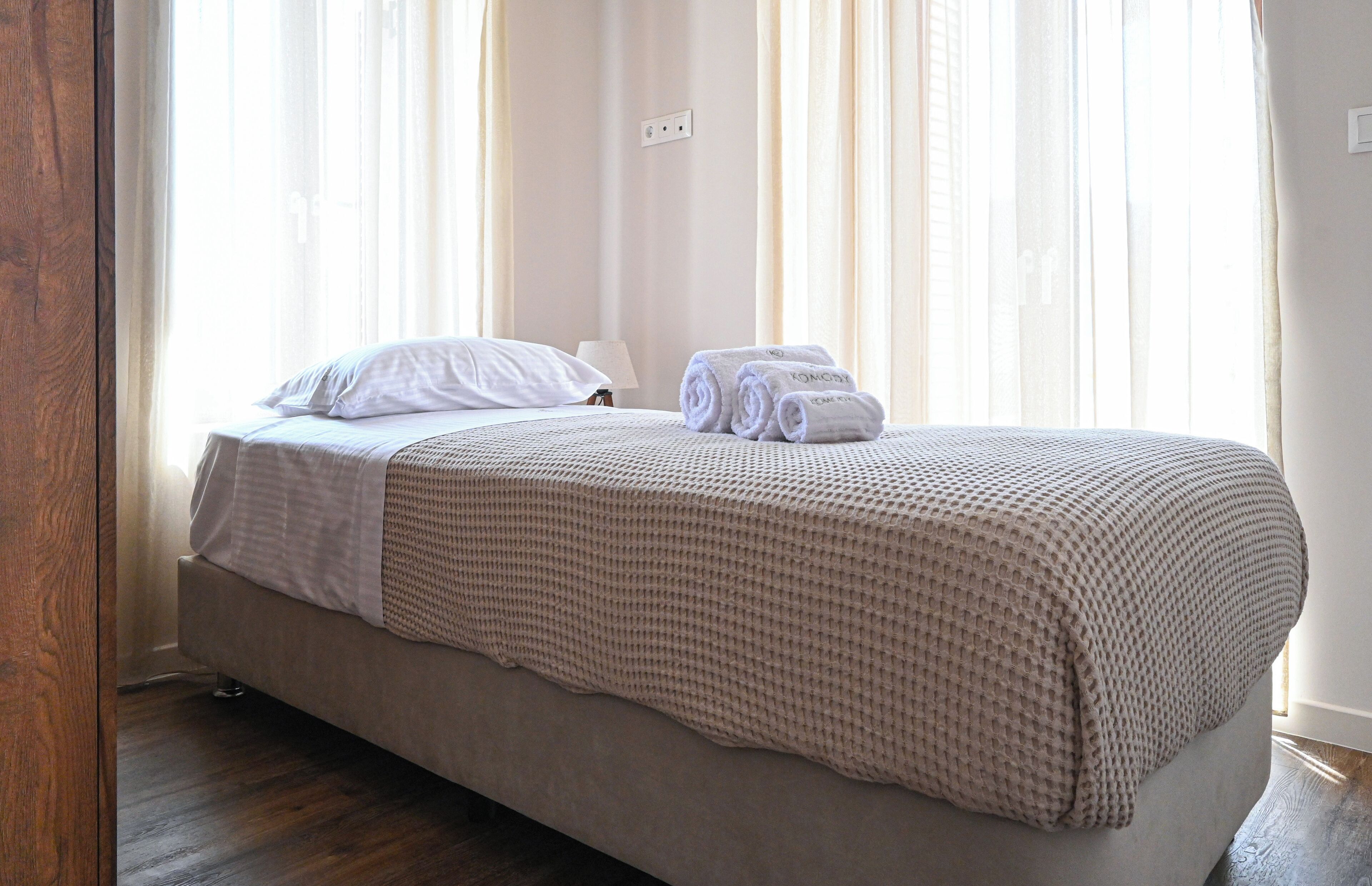 Una cama bien hecha con un edredón texturizado, una almohada y toallas enrolladas sobre un piso de madera.
