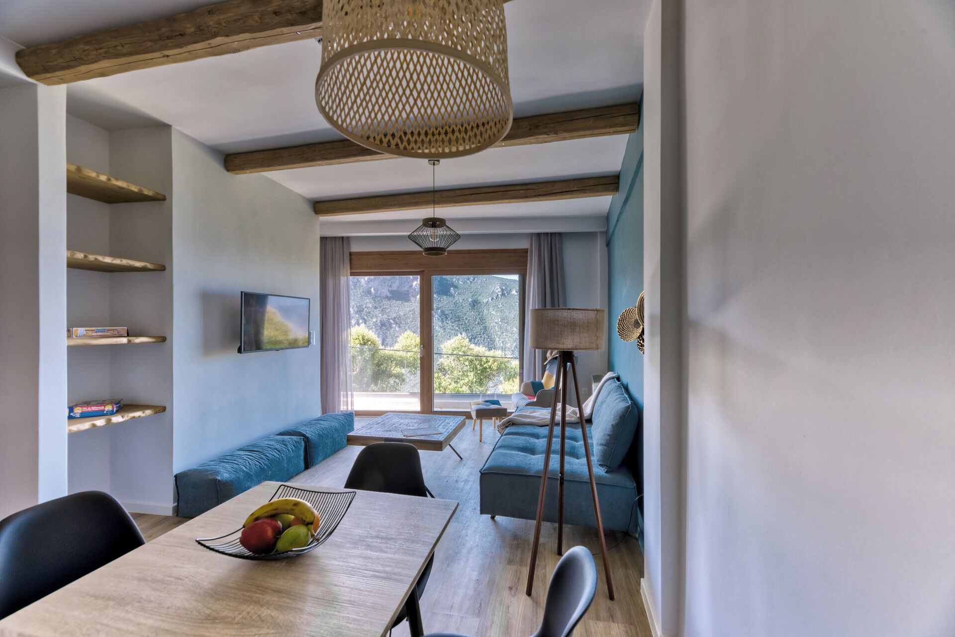 A modern living room with a wooden dining table, blue sofa, and a large window offering a view of the outdoors.