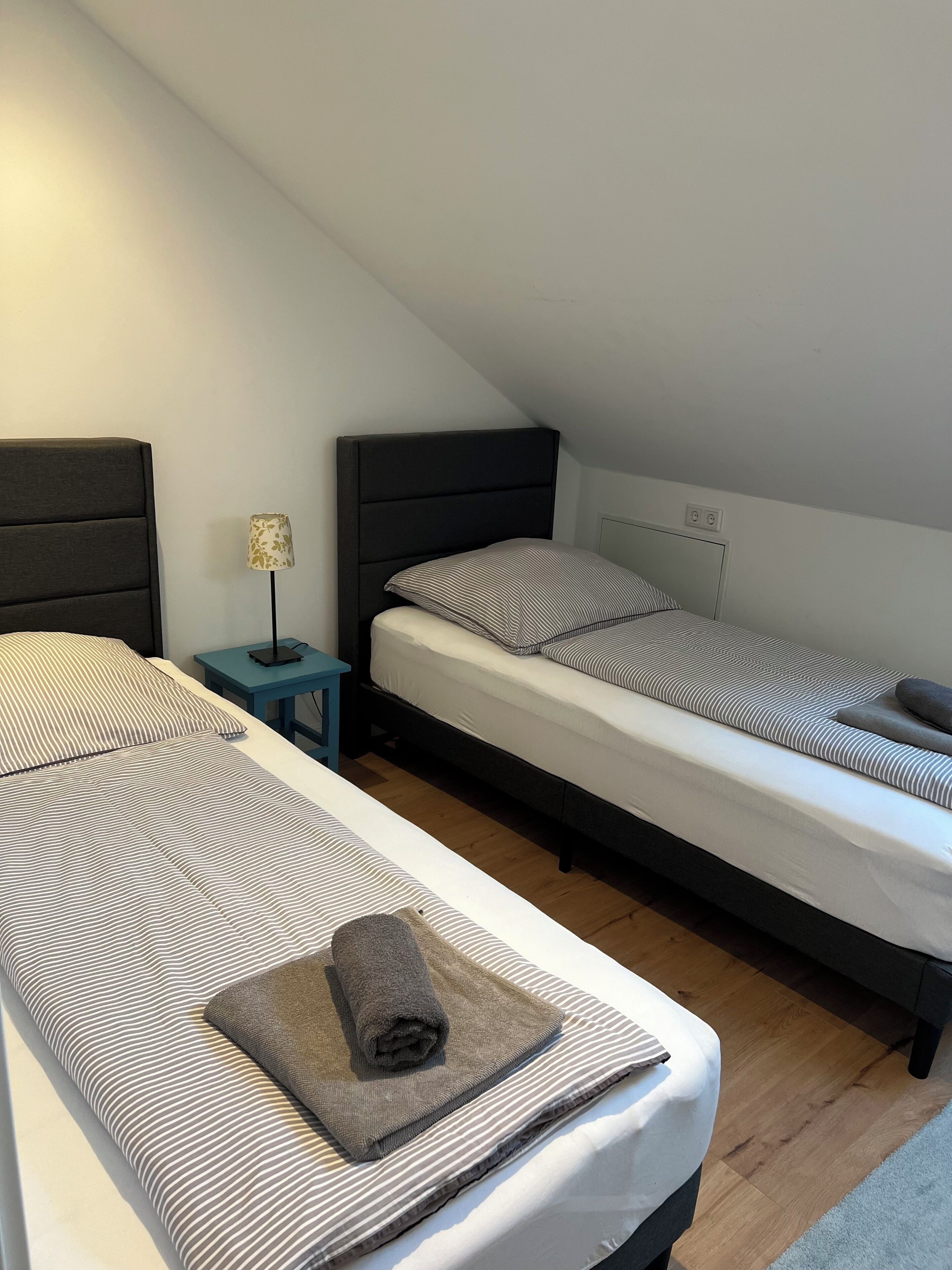 A small, cozy room with two single beds, a nightstand with a lamp, and a wooden floor.