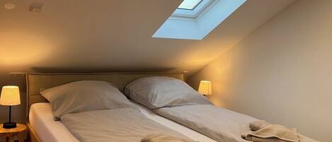 A bedroom with a skylight, a bed with pillows, a nightstand with a lamp, and a wooden floor.