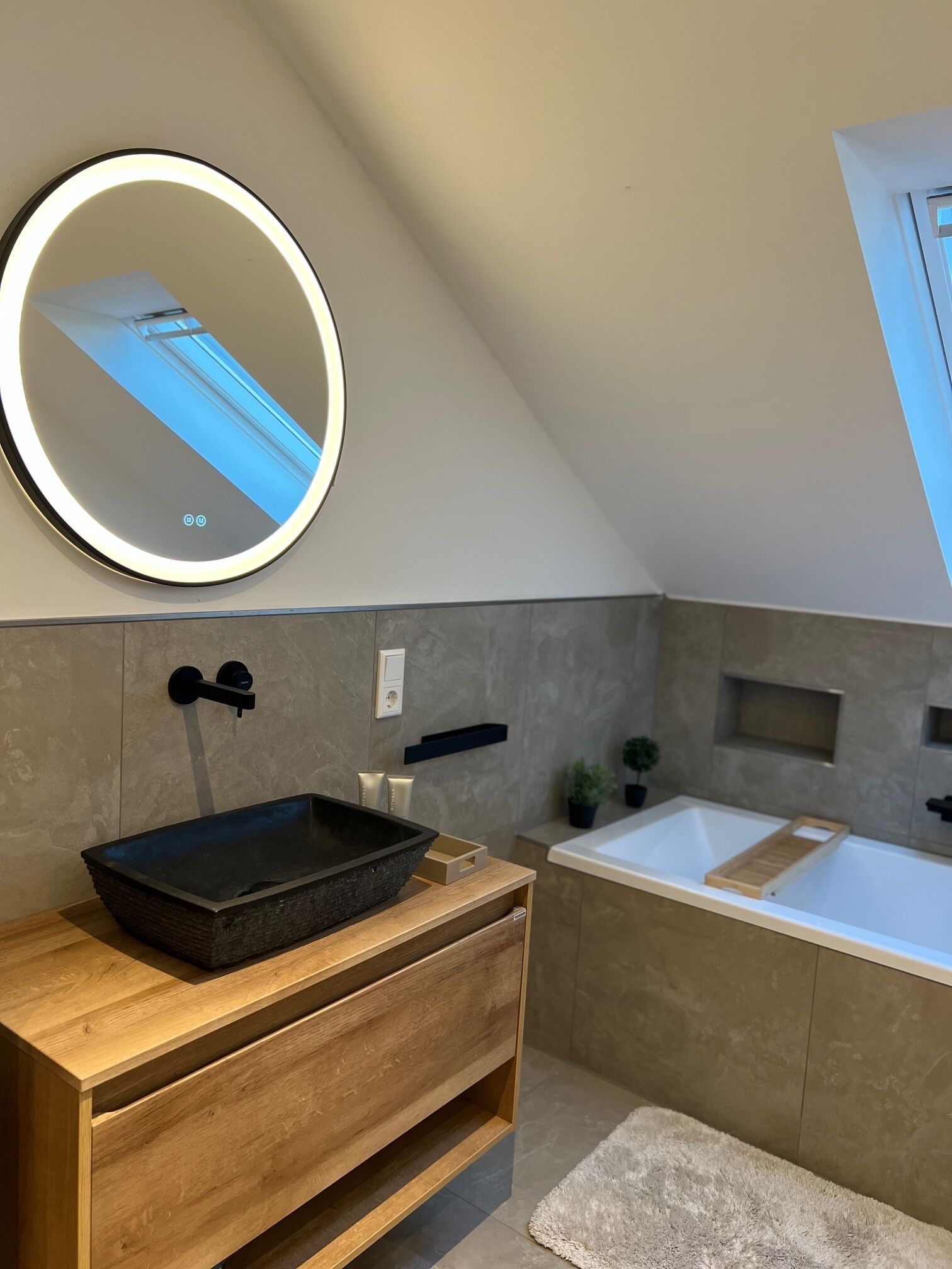 A modern bathroom with a wooden vanity, a black vessel sink, a white bathtub, and a round mirror with a light.