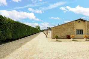 A gravel driveway leading to a row of stone buildings with a neatly trimmed hedge on one side.