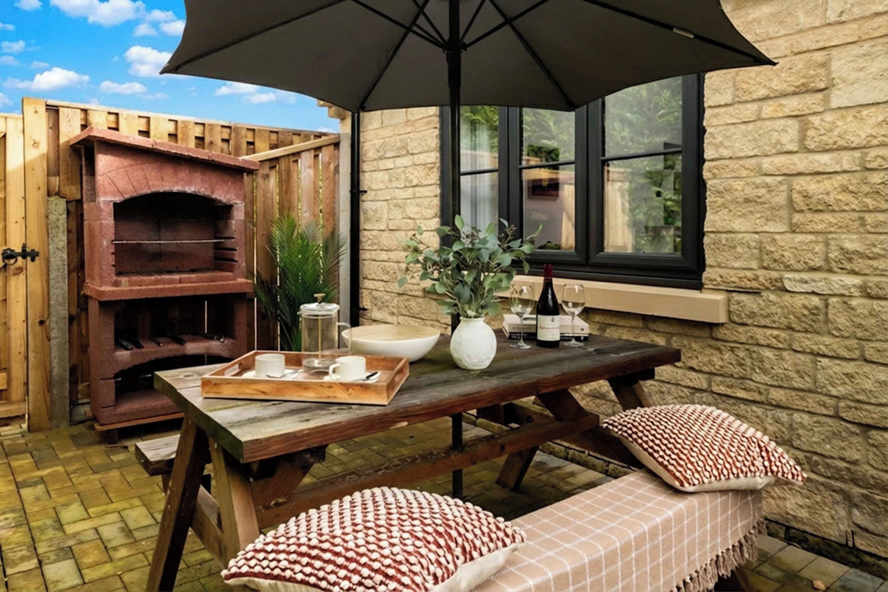 An outdoor patio with a stone wall, a wooden table, a pizza oven, and a large umbrella.