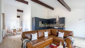 A modern living room with a brown leather sectional sofa, a dining area with a glass table, and a kitchen with dark cabinets and stainless steel appliances.
