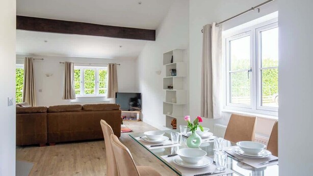 A modern living room with a glass dining table set for four, a brown sofa, and a TV in the background.