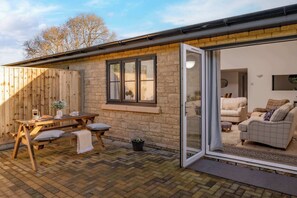 Une agréable terrasse extérieure avec une table et des bancs en bois, un mur en pierre et une porte-fenêtre coulissante donnant sur un espace de vie intérieur comprenant un canapé et un téléviseur.