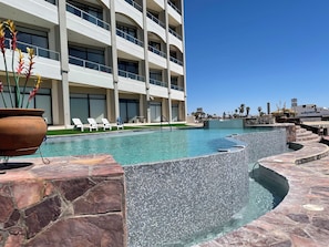 A pool area with a stone wall, lounge chairs, and a building with multiple balconies.