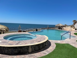 A swimming pool with a hot tub, surrounded by a stone deck and a grassy area, with a view of the ocean and beach amenities.