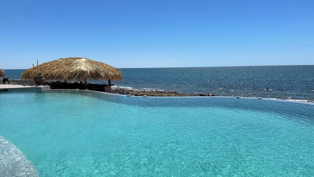 Ein klarer, blauer Infinity-Pool mit einer strohgedeckten Struktur und einer felsigen Küstenlinie.