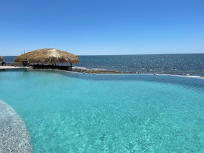 Une magnifique piscine à débordement d’un bleu profond, dotée d’une structure au toit de chaume et bordée d’un rivage rocheux.