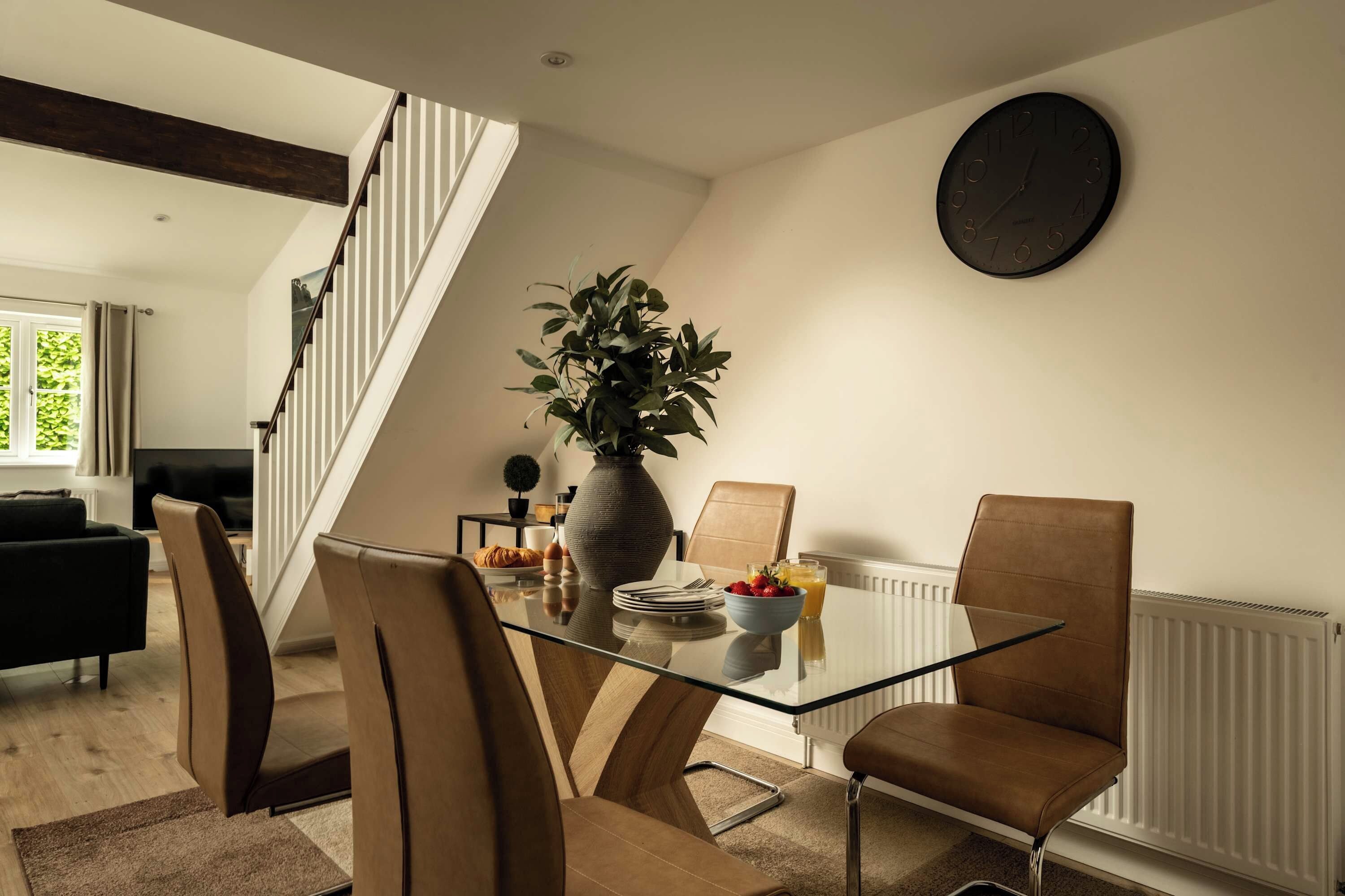 A dining area with a glass table, leather chairs, a vase with plants, and a wall clock.