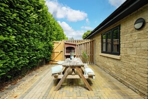 A wooden table with benches in a brick courtyard, surrounded by a hedge and a stone wall.