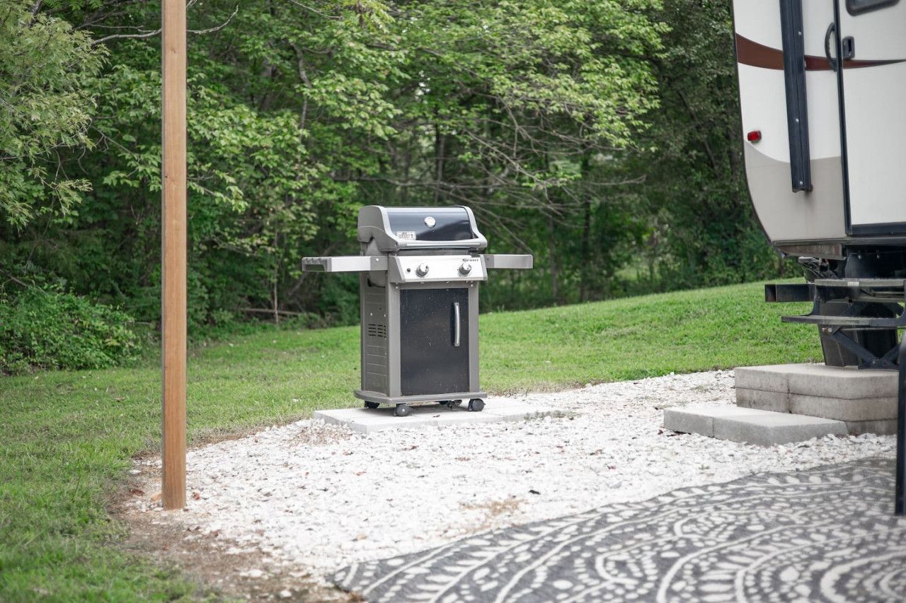 Un barbecue portatif doté d’un panneau de commande et d’un petit compartiment de rangement, placé sur une surface de gravier, à proximité d’un champ herbeux et d’un poteau en bois.