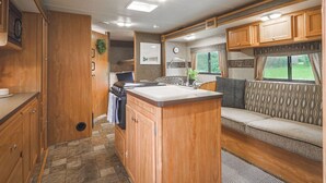 A compact RV kitchen with wooden cabinets, a central island, and a seating area with a patterned cushion.