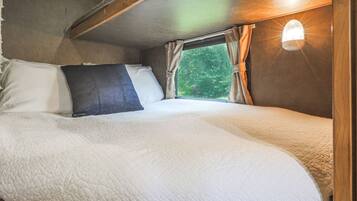 A bed with a white quilt and a dark pillow in a room with a window and a wall-mounted light.