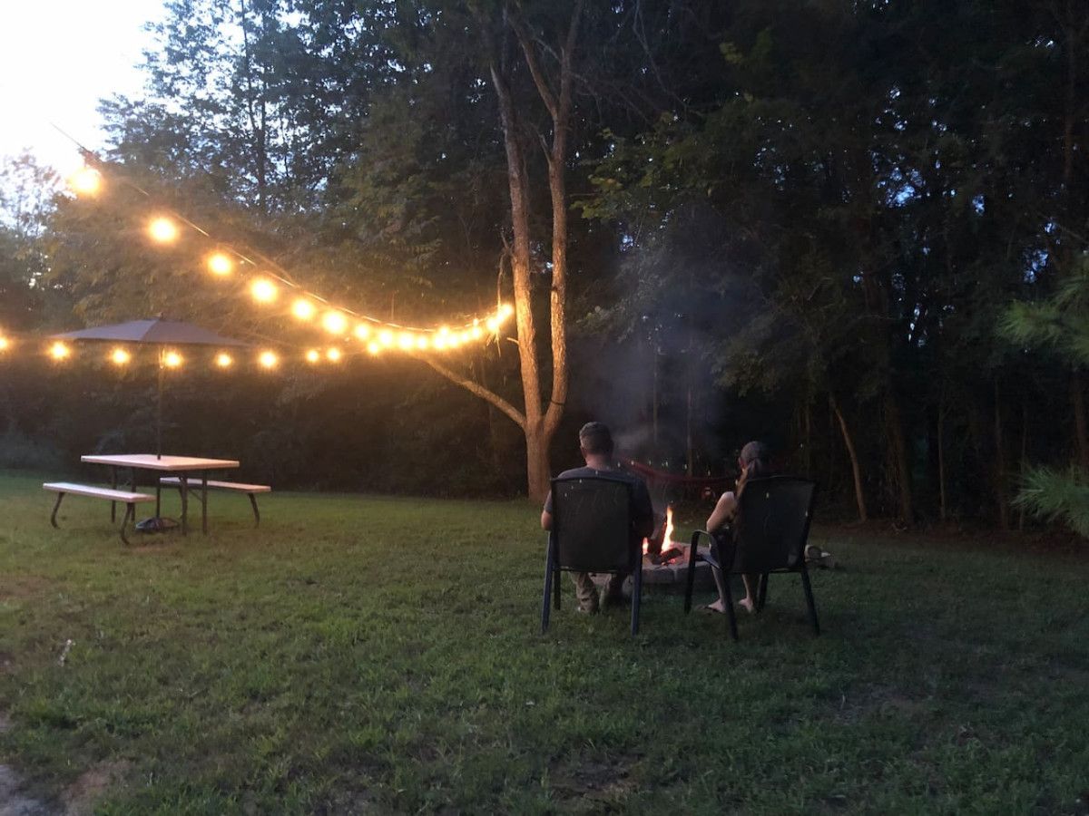 An outdoor setting with a picnic table and chairs, a campfire, and string lights.