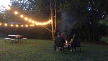 An outdoor setting with a picnic table and chairs, a campfire, and string lights.